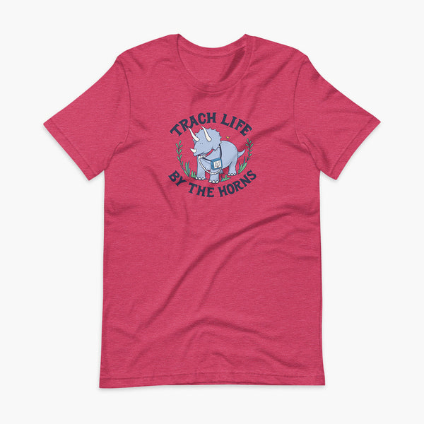 A happy triceratops dinosaur that has a trach or tracheostomy and a vent or ventillator to help him breathe. He also has a g-tube or gastronomy tube. There is text wrapping around the dinosaur that says Trach Life By The Horns. It is on a heather raspberry adult t-shirt.