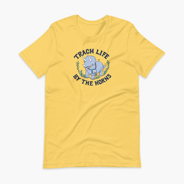 A happy triceratops dinosaur that has a trach or tracheostomy and a vent or ventillator to help him breathe. He also has a g-tube or gastronomy tube. There is text wrapping around the dinosaur that says Trach Life By The Horns. It is on a yellow adult t-shirt.