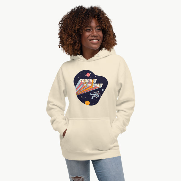 An illustration of an astronaut with a trach and tubing floating in outer space surrounded by stars and planets. Behind the astronaut, bold retro text reads “Trach It to the Limit,” with colorful light trails shooting from the words like a rocket launch. The design appears on a dark background. It is on a bone adult hoodie on a model.