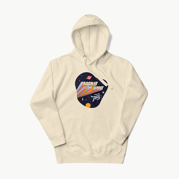 An illustration of an astronaut with a trach and tubing floating in outer space surrounded by stars and planets. Behind the astronaut, bold retro text reads “Trach It to the Limit,” with colorful light trails shooting from the words like a rocket launch. The design appears on a dark background. It is on a bone adult hoodie.