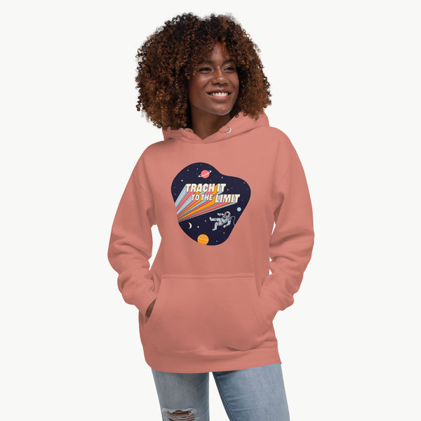 An illustration of an astronaut with a trach and tubing floating in outer space surrounded by stars and planets. Behind the astronaut, bold retro text reads “Trach It to the Limit,” with colorful light trails shooting from the words like a rocket launch. The design appears on a dark background. It is on a dusty rose adult hoodie on a model.
