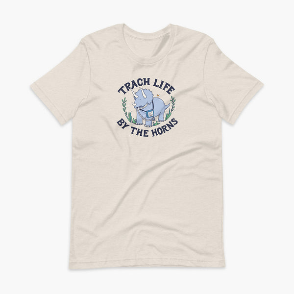 A happy triceratops dinosaur that has a trach or tracheostomy and a vent or ventillator to help him breathe. He also has a g-tube or gastronomy tube. There is text wrapping around the dinosaur that says Trach Life By The Horns. It is on a heather dust adult t-shirt.