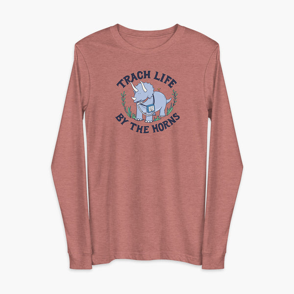 A happy triceratops dinosaur that has a trach or tracheostomy and a vent or ventillator to help him breathe. He also has a g-tube or gastronomy tube. There is text wrapping around the dinosaur that says Trach Life By The Horns. It is on a heather mauve blue adult long sleeve t-shirt.