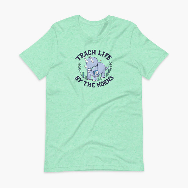A happy triceratops dinosaur that has a trach or tracheostomy and a vent or ventillator to help him breathe. He also has a g-tube or gastronomy tube. There is text wrapping around the dinosaur that says Trach Life By The Horns. It is on a heather mint adult t-shirt.