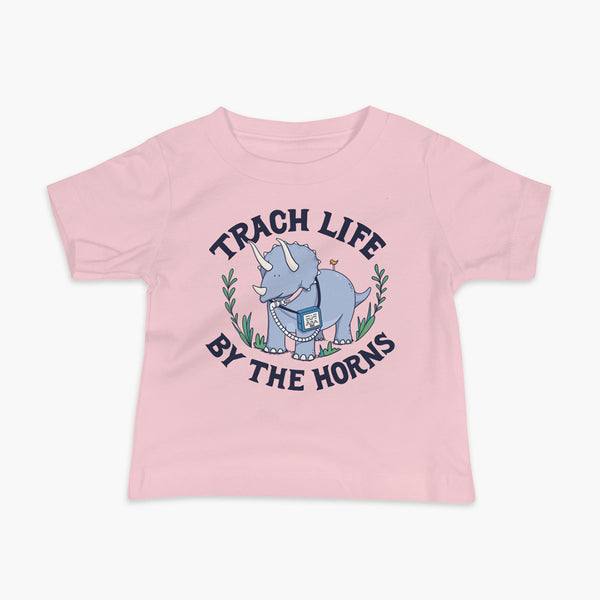 A happy triceratops dinosaur that has a trach or tracheostomy and a vent or ventillator to help him breathe. He also has a g-tube or gastronomy tube. There is text wrapping around the dinosaur that says Trach Life By The Horns. It is on a pink infant t-shirt.