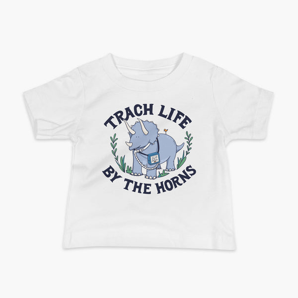 A happy triceratops dinosaur that has a trach or tracheostomy and a vent or ventillator to help him breathe. He also has a g-tube or gastronomy tube. There is text wrapping around the dinosaur that says Trach Life By The Horns. It is on a white infant t-shirt.