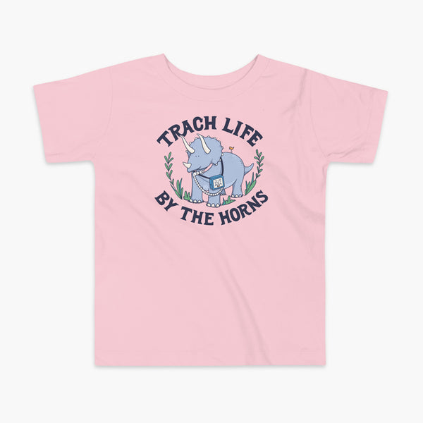 A happy triceratops dinosaur that has a trach or tracheostomy and a vent or ventillator to help him breathe. He also has a g-tube or gastronomy tube. There is text wrapping around the dinosaur that says Trach Life By The Horns. It is on a pink kids t-shirt.
