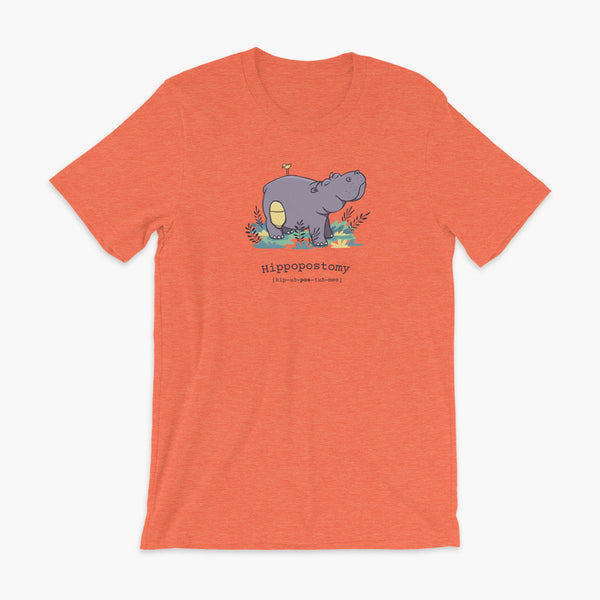 A Hippo or Hippopotamus with an ostomy bag — also known as a Hippopostomy. He is standing in some foliage smiling and has a bird on his back on a heather orange adult t-shirt.