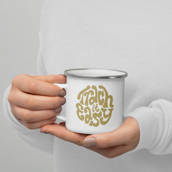 A white enamel mug with a white coating and silver rim with the text "Trach it Easy" in gold hand lettering on the side being held in someones hand.