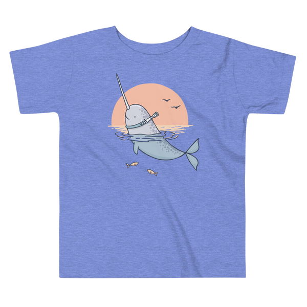A Narwhal that has a trach or tracheostomy pokes his head and horn through the water in front of a setting sun. He has a naturally built in stoma. It is on a heather columbia blue kids t-shirt.